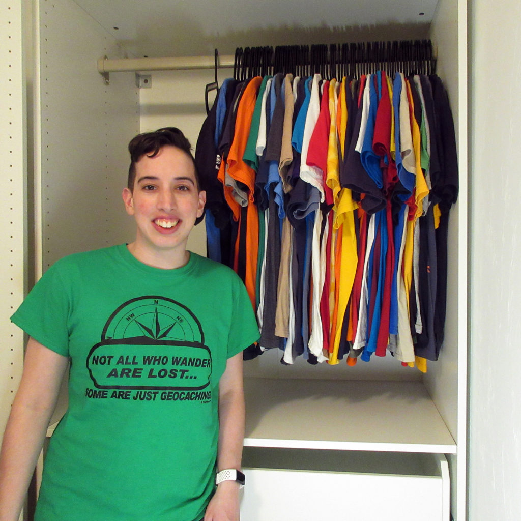 A person stands in front of a closet where 72 shirts hang with one hanger to the left empty. The person is wearing a green shirt with a design on it. The design features the top of a compass rose and under it the phrase "Note all who wander are lost... some are just geocaching."